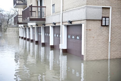 Residential building with multiple garage doors submerged in floodwaters reflecting a clear sky and surrounding trees, highlighting the need for Water Damage Restoration.